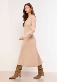 Beżowa sukienka dzianinowa z prążkowanym wzorem, z wysokim kołnierzem i wzorem warkoczowym. Długie rękawy i rozkloszowany dół. Zestawiona z brązowymi kozakami za kolano.