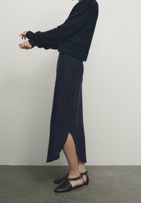 Tummansininen neulepaita pitkillä hihoilla, yhdistettynä tummansiniseen midi-hameeseen, jossa on sivureikä. Mustat matalat kengät täydentävät asua.