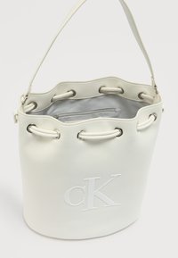 Λευκή τσάντα-κούπα με έναν λουρί, κορδόνι στο πάνω μέρος, ασημένιες οπές, εσωτερική τσέπη με φερμουάρ και ανάγλυφο λογότυπο "CK" στο μπροστινό μέρος.