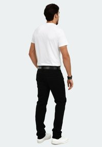 Schwarze Jeans mit slim Fit, kombiniert mit einem weißen T-Shirt mit kurzen Ärmeln und weißen Turnschuhen. Ein schwarzer Gürtel setzt einen Kontrast. Einfarbiger Hintergrund.