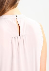 Top senza maniche di un delicato rosa chiaro con dettaglio a occhiello sulla schiena, tessuto morbido con pieghe sottili e una piccola chiusura a bottone nera sul collo.
