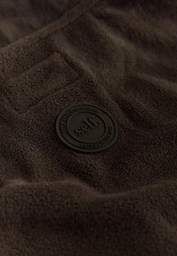 Brown fleece fabric with a plush texture, featuring a circular black rubber badge with "self. SELF ATHLETICS CLUB ESTABLISHED 2025."