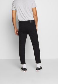 Svarta slim-fit jeans med medelhög midja, med bakfickor och ett lädermärke vid midjan, tillsammans med en grå T-shirt och sneakers.