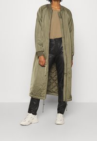 Veste bomber longue olive verte avec fermeture éclair, doublure matelassée, poignets côtelés, associée à un pantalon en similicuir noir et des baskets blanches.