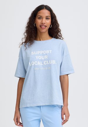Jeune femme aux cheveux bouclés portant un t-shirt bleu clair et un pantalon, le texte sur le t-shirt dit « Soutenez votre club local. Club de l'amour de soi. »