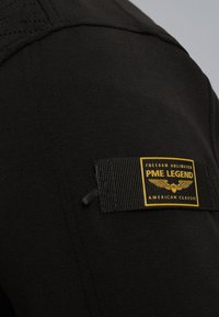 Camisa de algodón negra con un parche texturizado que presenta un logo amarillo de PME Legend y texto. El diseño incluye un cuello redondo y detalles acanalados.