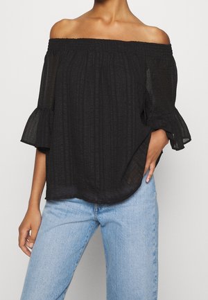 Top nero off-the-shoulder in tessuto leggero con strisce verticali testurizzate e maniche a campana, abbinato a jeans azzurri chiari.