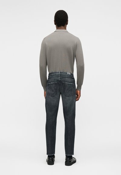 Άνδρας που φοράει γκρι μακρυμάνικο πουκάμισο, σκουρόχρωμο ξεβαμμένο τζην με τσέπες με πατς και μαύρα παπούτσια, στέκεται γυρισμένος με την πλάτη προς το μέρος μας σε απλό φόντο.