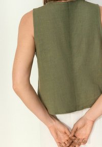 Top verde senza maniche con taglio dritto, realizzato in tessuto strutturato. La parte posteriore presenta una cucitura centrale, con le mani tenute dietro la schiena.