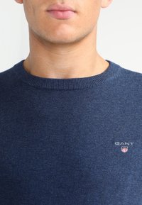 Pull bleu foncé en tricot avec un col rond. Présente un petit logo GANT brodé sur la poitrine. Texture lisse.