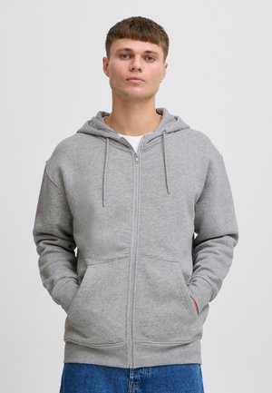 Ung mand iført en lysegrå hættetrøje med lynlås, hætte og forlommer, kombineret med blå jeans, stående mod en ensfarvet baggrund.