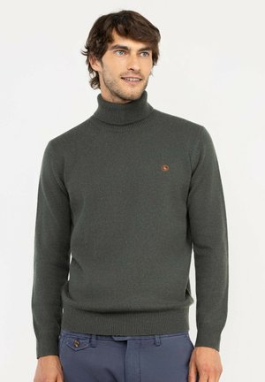 Hombre vestido con jersey de cuello alto gris oscuro con pequeño logo en el pecho y pantalones azules, de pie y mirando ligeramente hacia un lado.