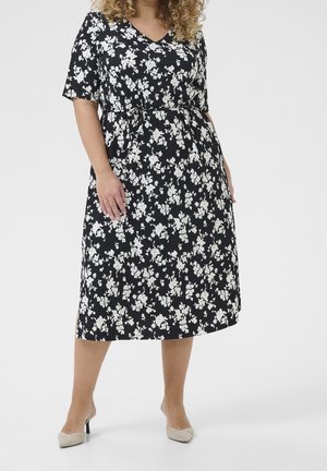 Frau trägt ein schwarzes Midi-Kleid mit weißem Blumenmuster, kurzen Ärmeln, V-Ausschnitt, gebundener Taille und beigen Absatzschuhen vor einfarbigem Hintergrund.