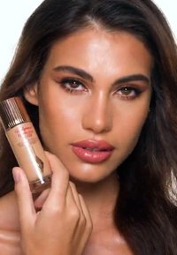 Foundation bottle with a clear base and gold-tone cap, held by a model. The liquid is a light beige shade, showcasing smooth texture.