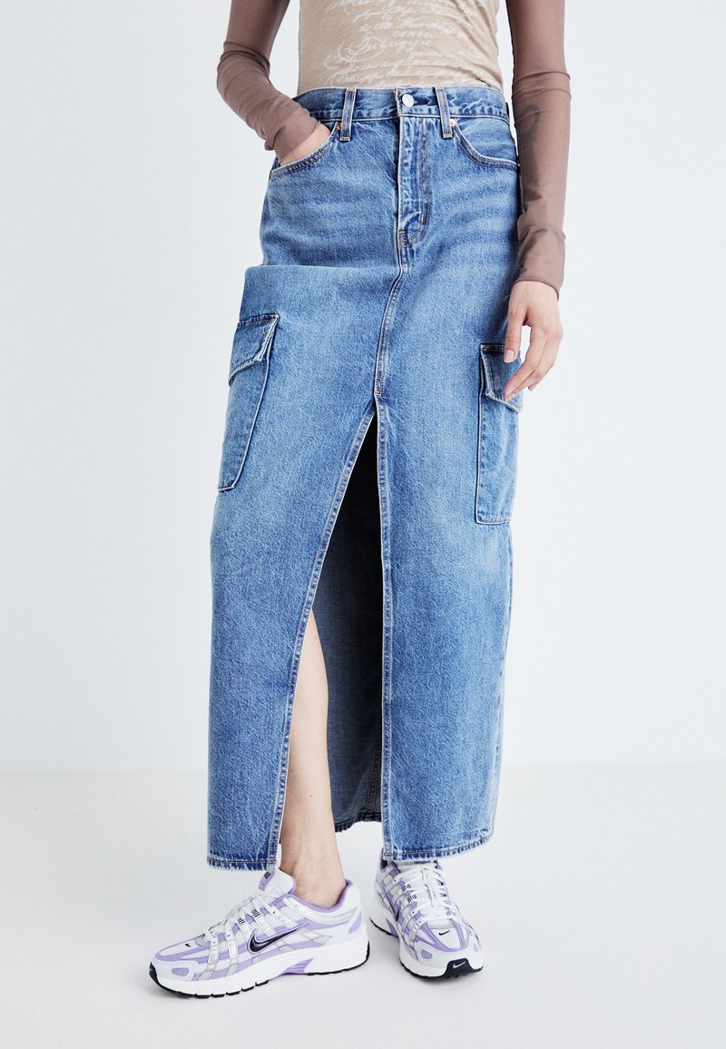 Personne portant une jupe en denim bleue avec une fente frontale et de grandes poches latérales, associée à des baskets violet clair et blanches.