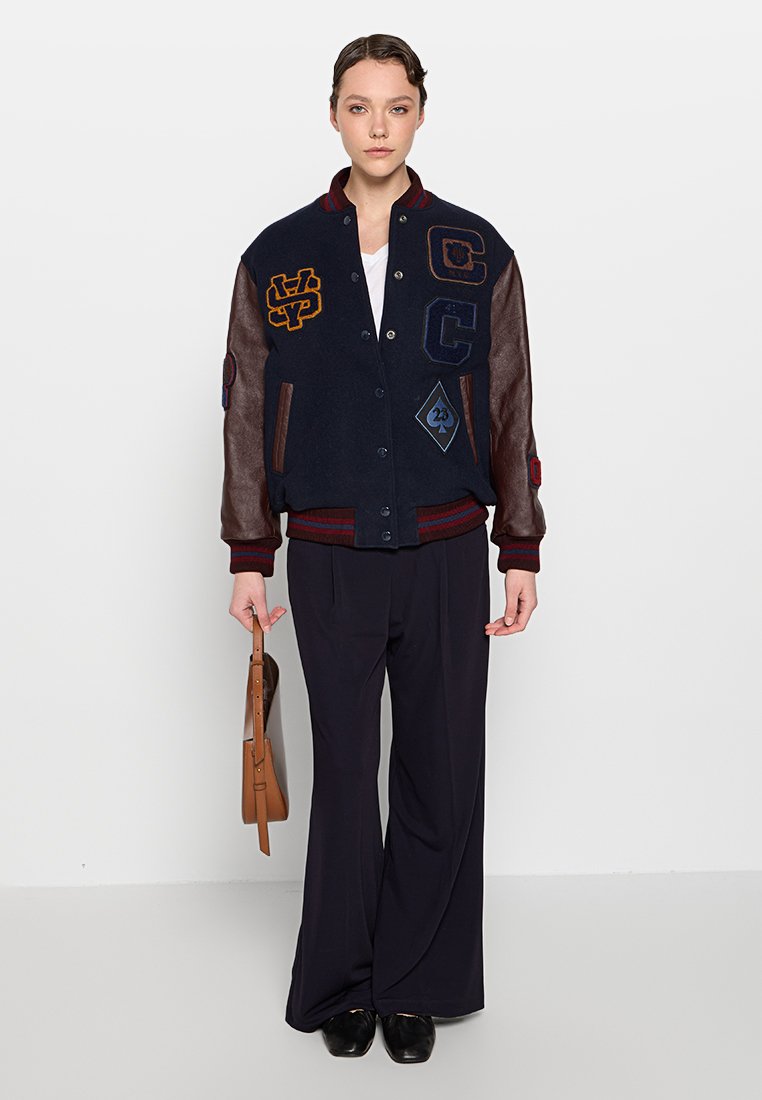 Donna che indossa una giacca varsity blu navy con maniche in pelle, pantaloni neri a gamba larga, scarpe nere e tiene una borsa marrone al suo fianco.