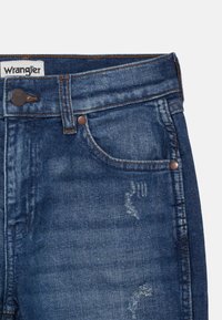Jeans de mezclilla azul oscuro con un lavado descolorido, que cuentan con herrajes de cobre, cinco bolsillos y un sutil envejecido cerca de la parte superior del muslo.