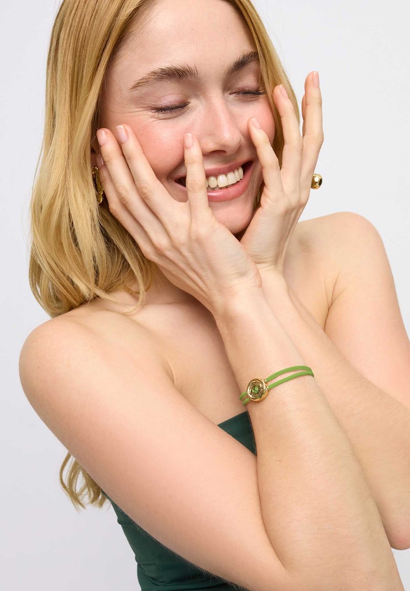 Le bracelet en caoutchouc vert est orné d'un motif de fleur dorée avec une pierre centrale. Le mannequin le porte avec une tenue sans bretelles de couleur vert foncé.
