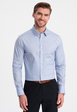 Camisa de manga larga azul claro con un pequeño patrón de puntos, cuello abotonado y botones blancos. Llevada con pantalones negros y un cinturón marrón.