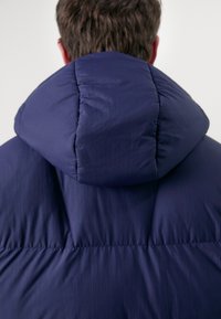 Doudoune bleu marine avec col montant et capuche, fabriquée en tissu texturé avec un matelassage horizontal et sans quincaillerie visible.