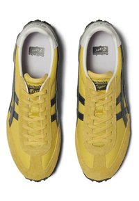 Scarpe atletiche gialle con accenti neri, dotate di una tomaia in camoscio e mesh, punta rotonda e suola testurizzata. Lacci piatti e dettagli con logo visibili.