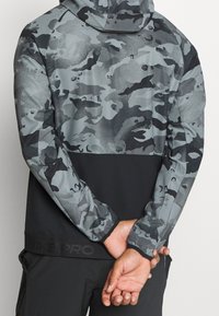 Kapsáčová bunda s šedým maskáčovým vzorem, s černým spodním panelem a značkováním Nike Pro na lemu. Hladká látka s volným střihem.