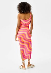 Šaty dokonale obepínající postavu v živé růžové a oranžové vlnité vzoru, s designem jednoho ramene a bočním rozparkem. Doplněné krémovými sandály na platformě.