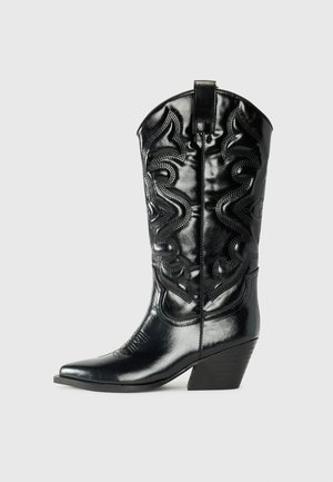 Bottine cowboy en cuir noir avec un bout pointu, des coutures décoratives et un talon incliné. Présente une texture lisse et des reliefs.