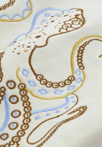 Vyšívaný dizajn chobotnice sohnedými, modrými a žltými niťami na svetlom krémovom plátne, s detailnými vírmi a kruhovými vzormi.