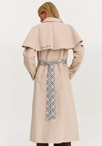 Žena v béžovém trenčkotu s vzorovaným páskem a kápí, stojící s rukama na bocích, obrácená zády k divákovi.
