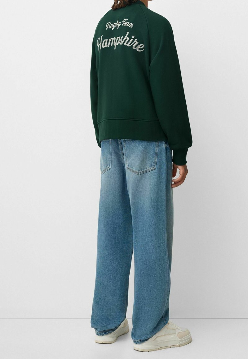 Sudadera verde con el texto blanco "Equipo de Rugby Hampshire", combinada con jeans de mezclilla azul sueltos y zapatillas de color claro. Vista trasera mostrada.