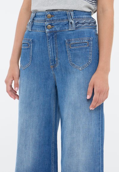 Modré džínové kalhoty širokého střihu s vysokým pasem na dvojité knoflíky, detail propleteného pásku a předními kapsami. Hladká textura s jemným vyblednutím.