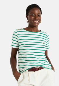 Camiseta de manga corta a rayas verdes y blancas con tres botones dorados en el hombro y un logo bordado, combinada con pantalones de color claro.