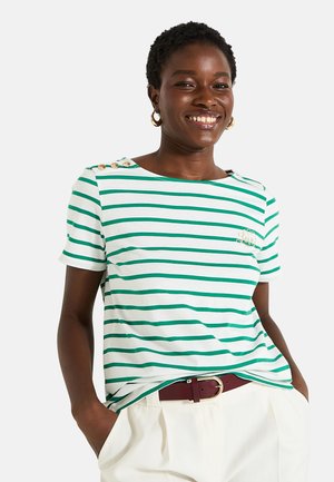 Camiseta de manga corta a rayas verdes y blancas con tres botones dorados en el hombro y un logo bordado, combinada con pantalones de color claro.