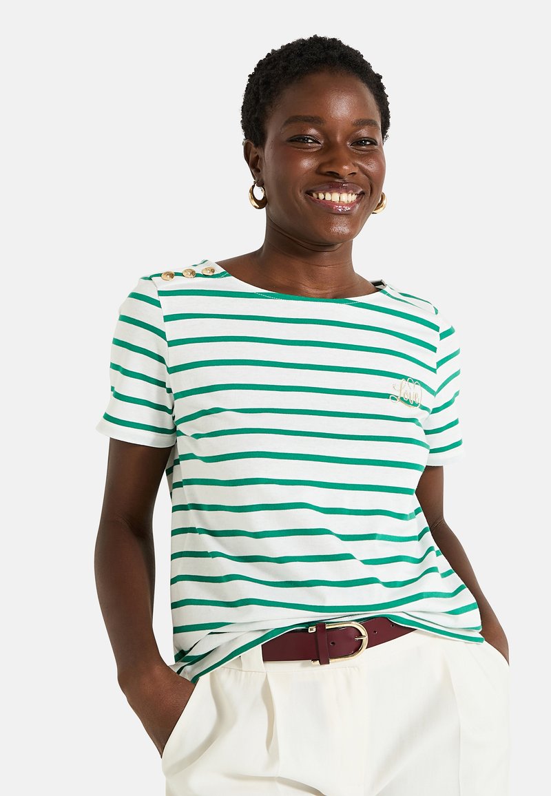 Camiseta de manga corta a rayas verdes y blancas con tres botones dorados en el hombro y un logo bordado, combinada con pantalones de color claro.