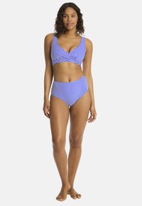 Donna in piedi a piedi nudi che indossa un costume da bagno a due pezzi blu e bianco con un top avvolgente e slip a vita alta.