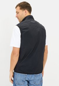Schwarze leichte Weste mit hochgeschlossenem Kragen, glatter Textur und einem Logo auf dem Rücken, getragen über einem weißen T-Shirt und blauen Jeans.
