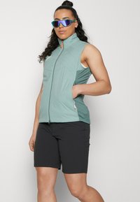 Světle modrá bezrukávová vesta se zipem na přední straně, se zpevněným designem a boční kapsou. Spárováno s černými, přiléhavými šortkami. Reflexní sluneční brýle.