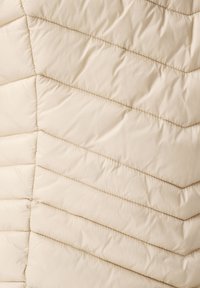 Veste matelassée en beige clair avec un motif en zigzag. Texture lisse avec des coutures horizontales et diagonales pour l'isolation.