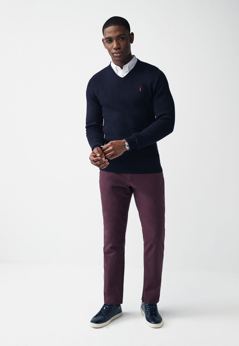 Marineblå strikket sweater med V-hals, parret med bordeaux chinos og mørke sneakers. Bemærkelsesværdig kontrast mellem sweateren og bukserne. Hvid skjorte med krave under.