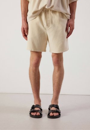Man die beige katoenen shorts met trekkoord draagt, gecombineerd met zwarte sandalen met gesp, staand op een lichtgrijze vloer tegen een witte achtergrond.