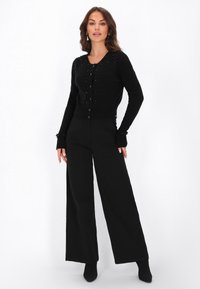 Juodas kardiganas su apvaliu iškirpimu, mygtukų uždarymu, puoštas tekstūruotais akcentais ant pečių, derinamas su plačiomis juodomis kelnėmis.