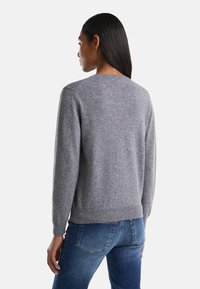 Šedý pletený svetr s kulatým výstřihem, žebrovanými manžetami a lemem. Nositý přes modré džíny, ukazující texturovanou tkaninu a uvolněný střih zezadu.