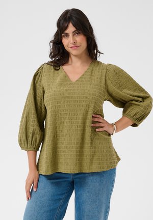 Vrouw met donker haar die een olijfgroene, getextureerde V-hals blouse met pofmouwen en een blauwe spijkerbroek draagt, poserend met hand op de heup.