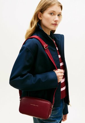 Junge Frau mit blondem Haar trägt eine marineblaue Jacke, einen gestreiften roten und pinken Pullover und eine burgunderrote Leder-Umhängetasche mit verstellbarem Riemen.