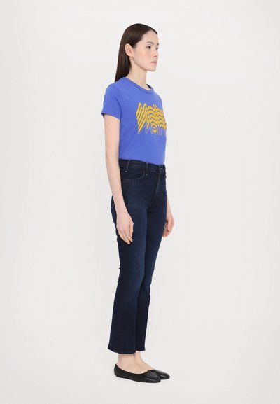 Blå grafisk t-skjorte med gul bølgemønster, kombinert med mørkeblå, tettsittende jeans og svarte flate sko. Modell står sidelengs.