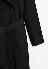 Mustat trench-takit, joissa on kaksirivinen malli, kaksi nappia, leveä vyö ja sileä kangas, jolla on raikas tekstuuri.