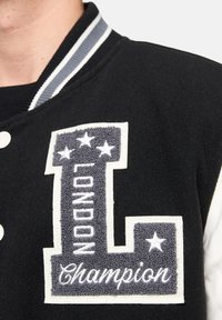 Chaqueta universitaria negra con textura de lana, parche bordado gris con "LONDRES" y estrellas, acentos blancos y un cuello a rayas.