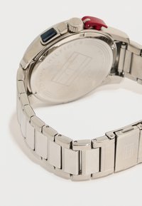 Ασημένιο μεταλλικό ρολόι χειρός Tommy Hilfiger με συνδεδεμένο λουρί, στρογγυλή πίσω θήκη και κόκκινα και μαύρα πλαϊνά κουμπιά σε λευκό φόντο.