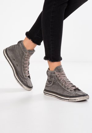 Personne portant des baskets montantes grises avec lacets et un jean noir à ourlet effiloché, debout sur une surface blanche.
